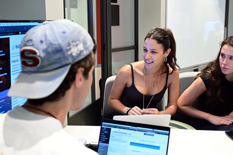 Three young people collaborate in an office discussing information on their laptops and monitors.