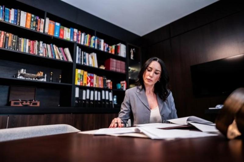 Woman in grey blazer working at a desk with papers, bookshelf in background.