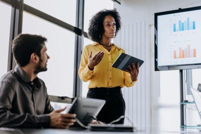 Businesswoman presenting data from a tablet on a large screen to a colleague.