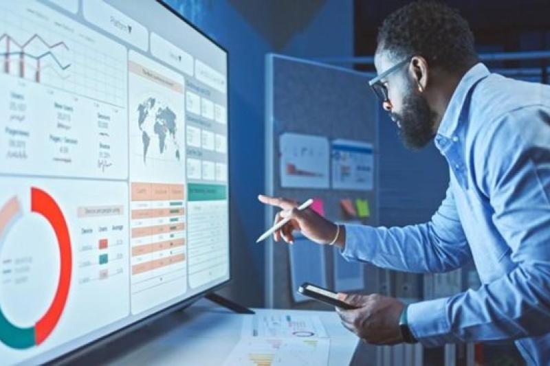 Man in blue shirt and glasses pointing at a large screen displaying data dashboards and charts.